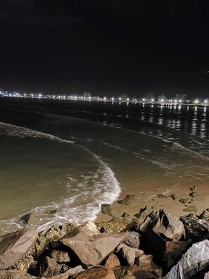 A nighttime beach scene with waves crashing onto rocks and a city skyline in the background. - Brisa e Mar Townhouse (Caraguatatuba)