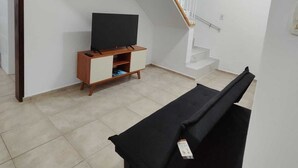 A modern living room with a flat-screen TV on a wooden cabinet, a black sofa, and a staircase with white railings. - Brisa e Mar Townhouse (Caraguatatuba)