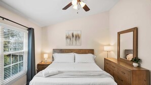 Una camera da letto con un letto, lampade sui comodini, una ventola a soffitto, una finestra con tende e uno specchio fissato alla parete.