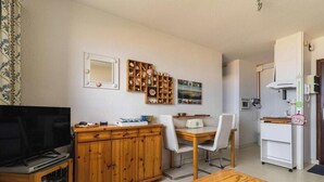 Ett kompakt bostadsutrymme med en TV, ett träskåp, ett matbord med stolar, en liten köksdel och en vägghängd hylla med dekorativa föremål.