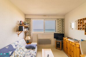 A room with a sofa, a coffee table, a TV, a bookshelf, and a window with a view of the sea. - 2-person apartment near the beach in Sables-d’Or-les-Pins | Ref 116 (SABLES D OR LES PINS)