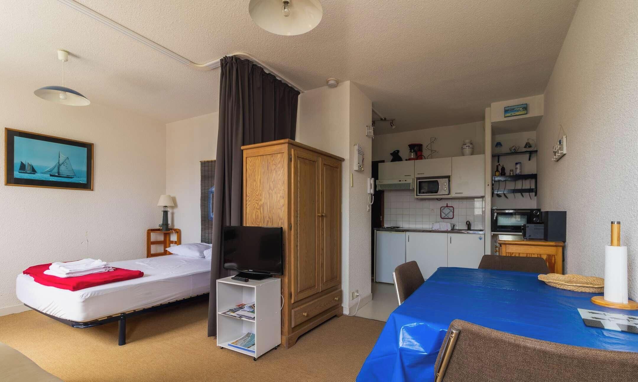 A small hotel room with two beds, a TV, a wooden wardrobe, a kitchenette, and a dining area.