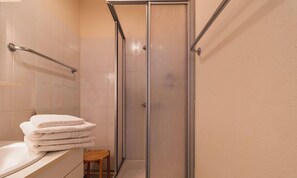 A bathroom with a glass shower enclosure, a stack of white towels, and a wooden stool. - 2-person apartment near the beach in Sables-d’Or-les-Pins | Ref 115 (SABLES D OR LES PINS)