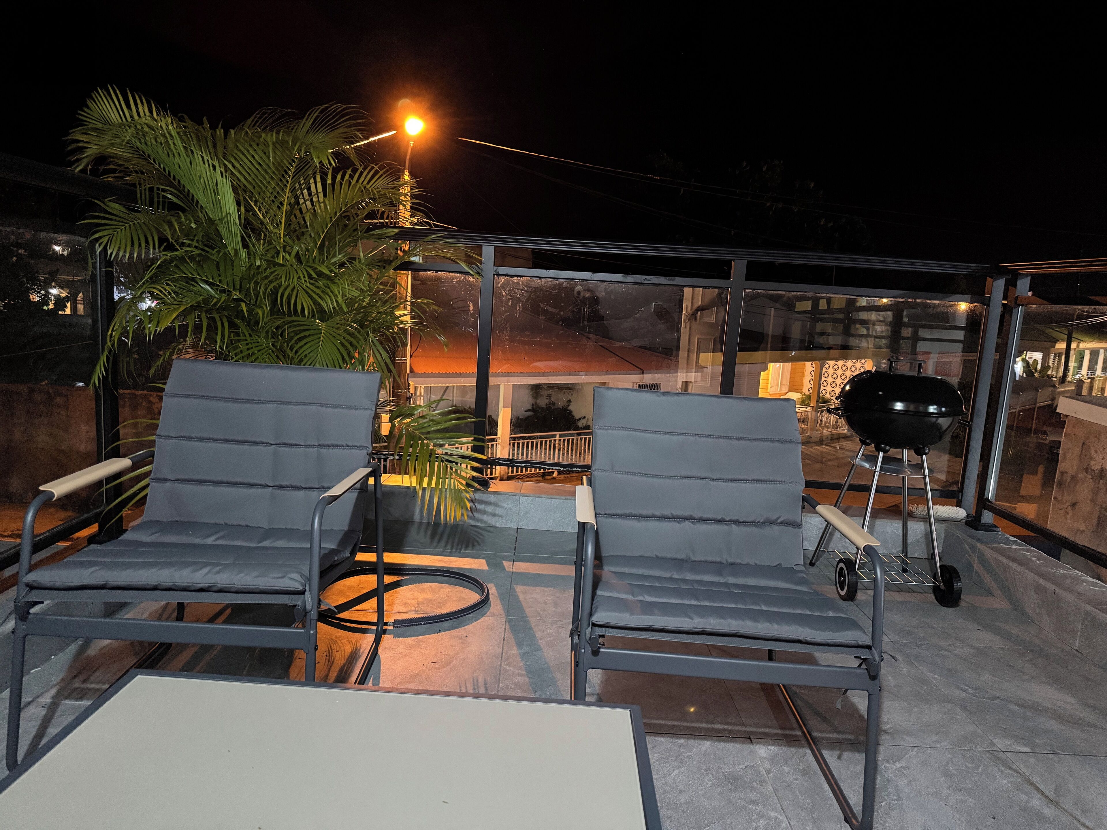 A rooftop terrace with two grey lounge chairs, a barbecue grill, and a glass enclosure.