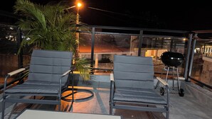 Terraza en la azotea con dos sillones grises, una parrilla y un cerramiento de vidrio.