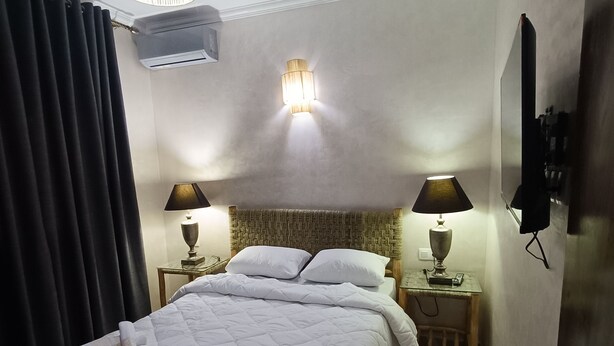 Een netjes opgemaakt bed met een witte dekbedovertrek, twee bedlampen, een aan de muur bevestigde televisie en een plafondlamp.