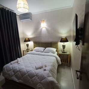 Een netjes opgemaakt bed met een witte dekbedovertrek, twee bedlampen, een aan de muur bevestigde televisie en een plafondlamp.