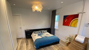 Un dormitorio con cama, mesita de noche, silla de mimbre, un cuadro y una lámpara de techo.