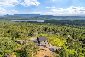 Ein Haus, umgeben von dichtem Wald, mit einem See und Bergen im Hintergrund.