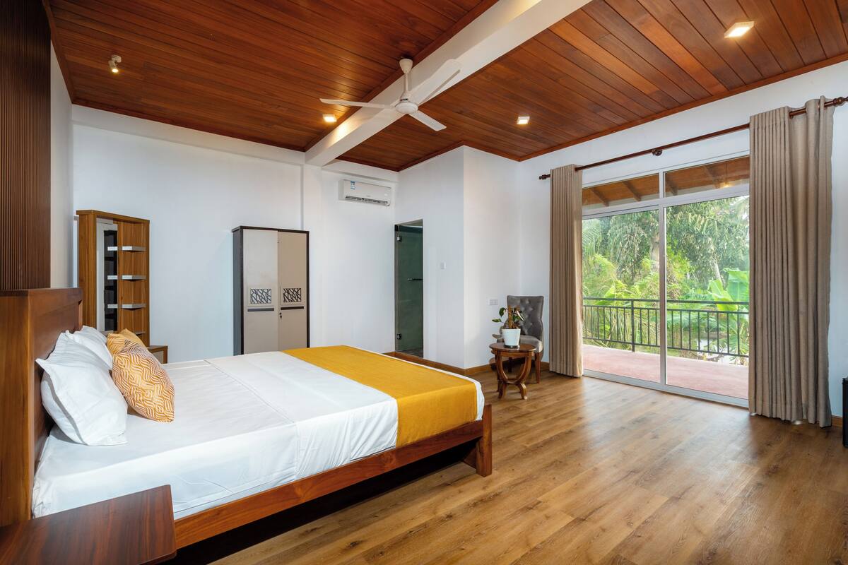 Une chambre avec un plafond en bois, un lit avec une literie blanche et jaune, un ventilateur de plafond, une fenêtre avec des rideaux et une vue sur la verdure à l’extérieur.