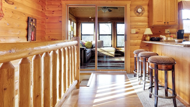 A wooden cabin interior with a balcony, a kitchen area, and a living room with a sofa and a window.