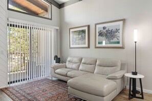 Un salon avec un canapé d’angle beige, un tapis à motifs, deux œuvres d’art encadrées, une lampe sur pied et une vue à travers une porte coulissante.