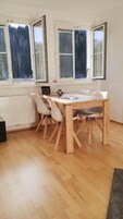 Un comedor con una mesa y sillas de madera, ventanas amplias con vistas a árboles y un radiador en la pared.