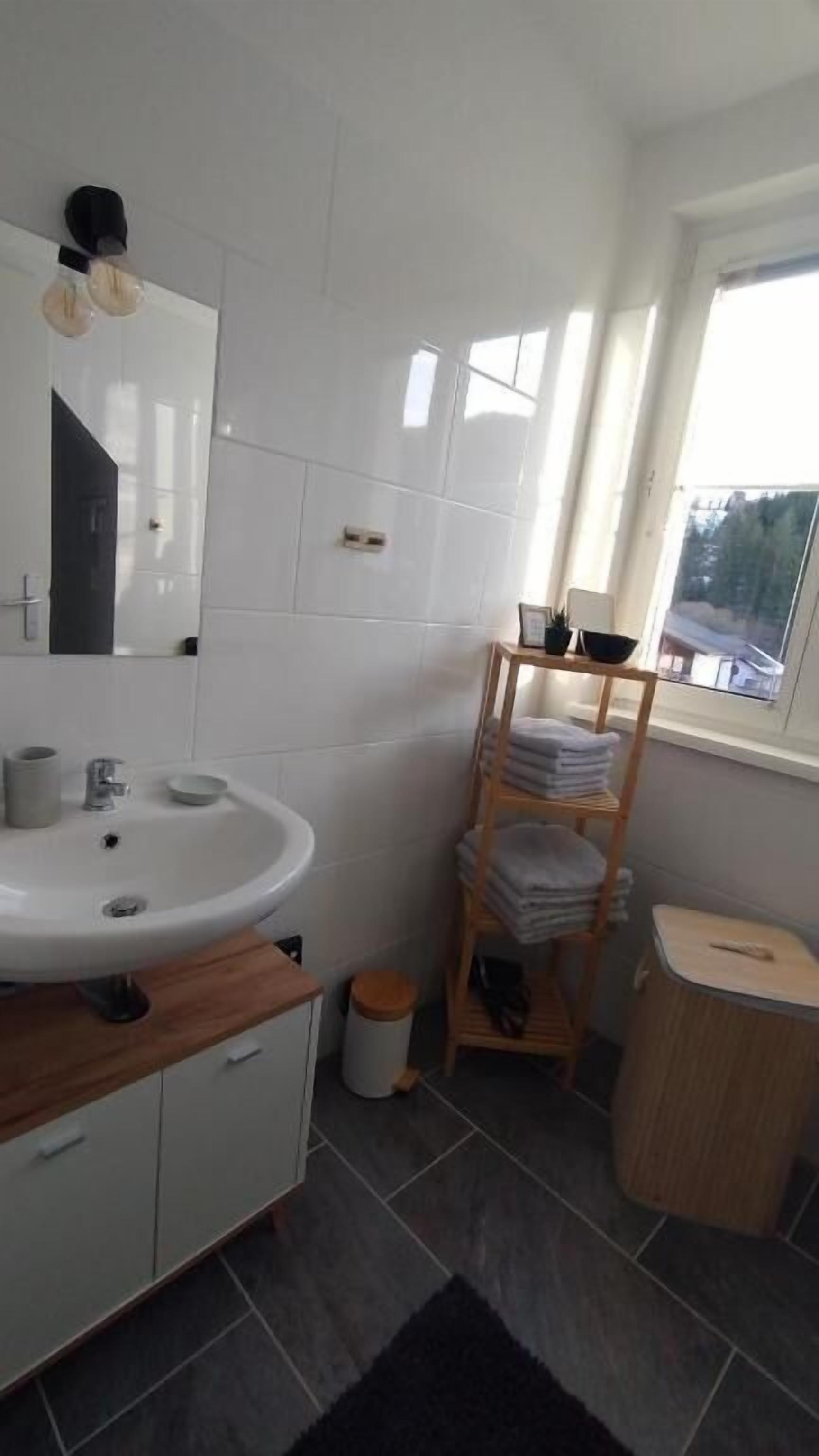 A modern bathroom with a white sink, mirror, and wooden vanity. There is a small window with a view of trees and a building outside.