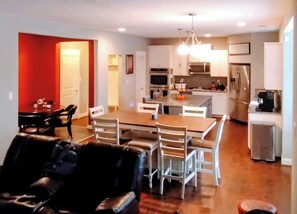 A modern kitchen with a central island, white cabinetry, and stainless steel appliances. There is a dining area with a wooden table and chairs.