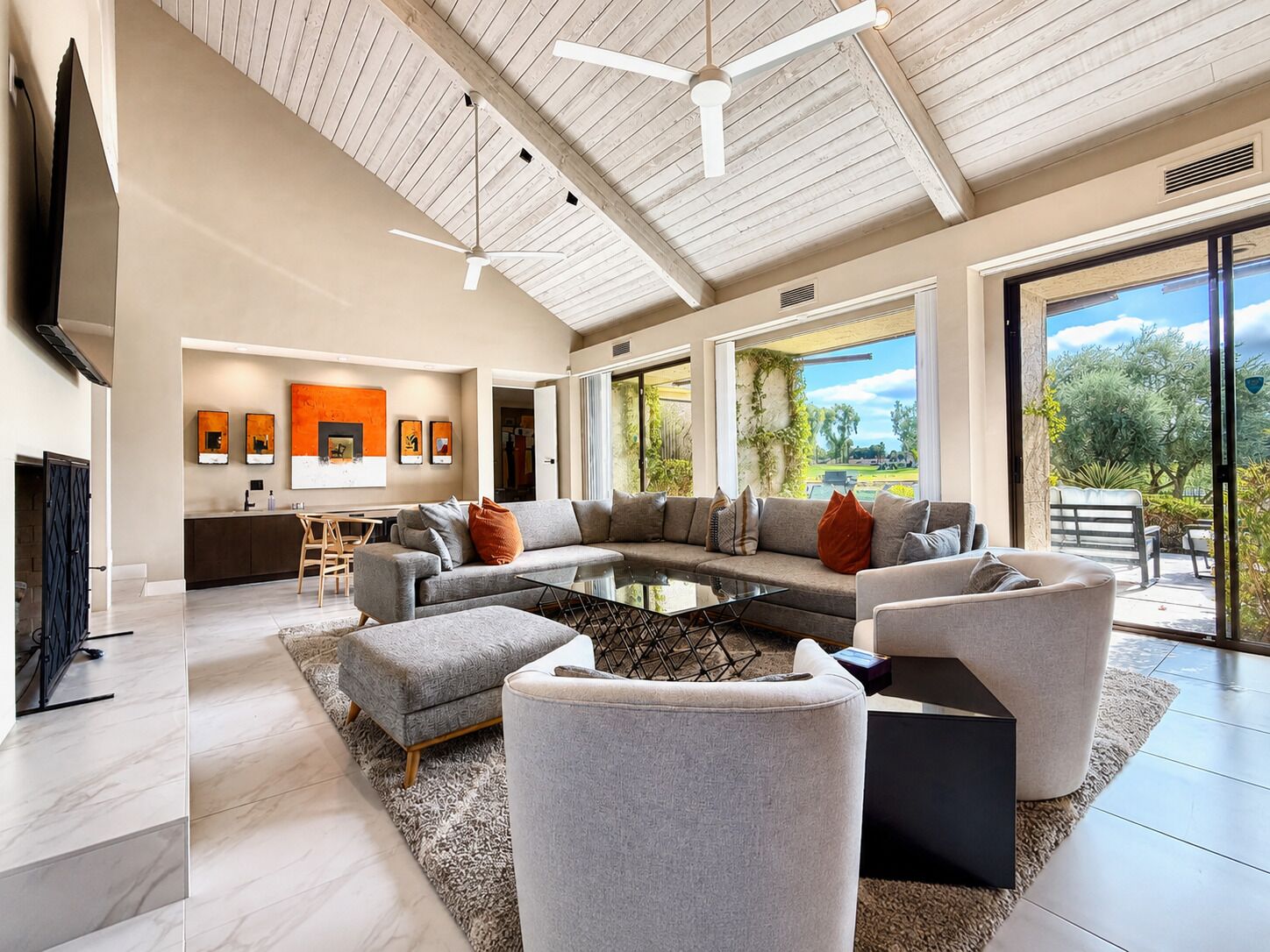 A spacious living area with a large sofa, a coffee table, and a TV. There are multiple ceiling fans and a view of the outdoors through large windows.