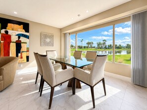 A dining area with a glass table and four chairs, a painting on the wall, and a view of a lake and golf course. - Grande Vista Villa: 3 bed/3 bath Luxury Living; Stunning Lake & Mountain Views | a Greenday property (Rancho Mirage)