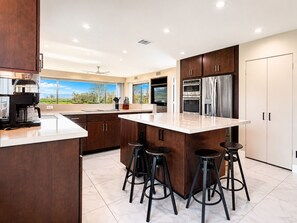 A modern kitchen with a large island, bar stools, stainless steel appliances, and a view of the outdoors. - Grande Vista Villa: 3 bed/3 bath Luxury Living; Stunning Lake & Mountain Views | a Greenday property (Rancho Mirage)