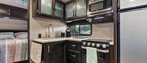 Cucina compatta per camper con fornello, microonde e finestra vista foresta.