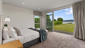 A bedroom with a large bed, a nightstand, a lamp, and a view of a garden and lake through a sliding glass door. - Lakefront Kuiwai - lakefront with spa pool (Taupo)