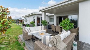 A modern outdoor living area with wicker furniture, a wooden deck, and a glass door leading to a covered patio. - Lakefront Kuiwai - lakefront with spa pool (Taupo)