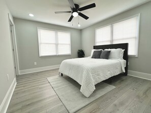 Une chambre à coucher avec un lit, un ventilateur de plafond et deux fenêtres équipées de stores.