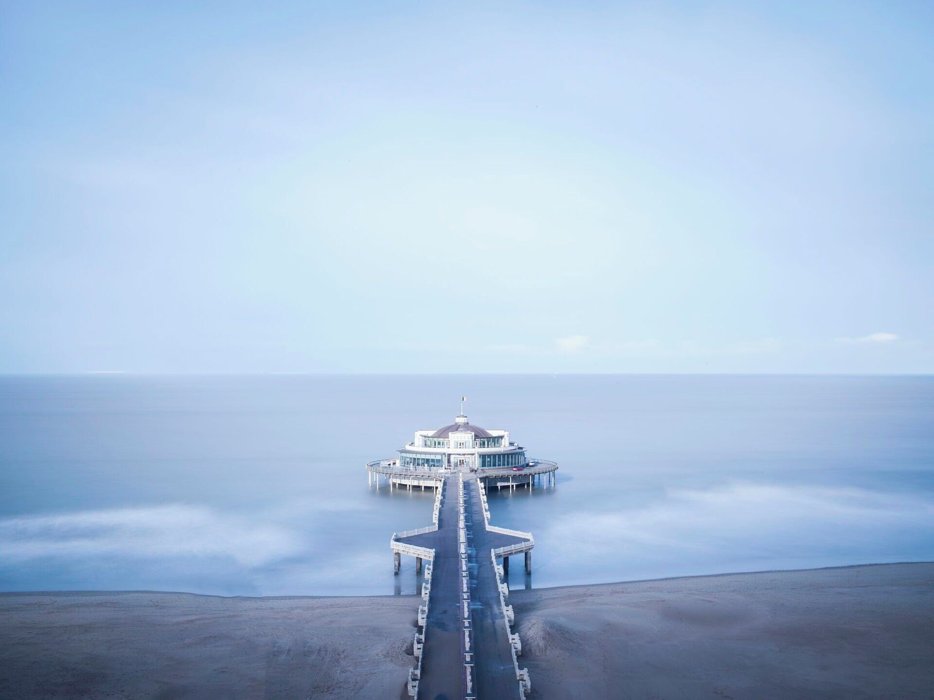 A pier leading to a building with a dome and multiple levels, extending into calm waters.