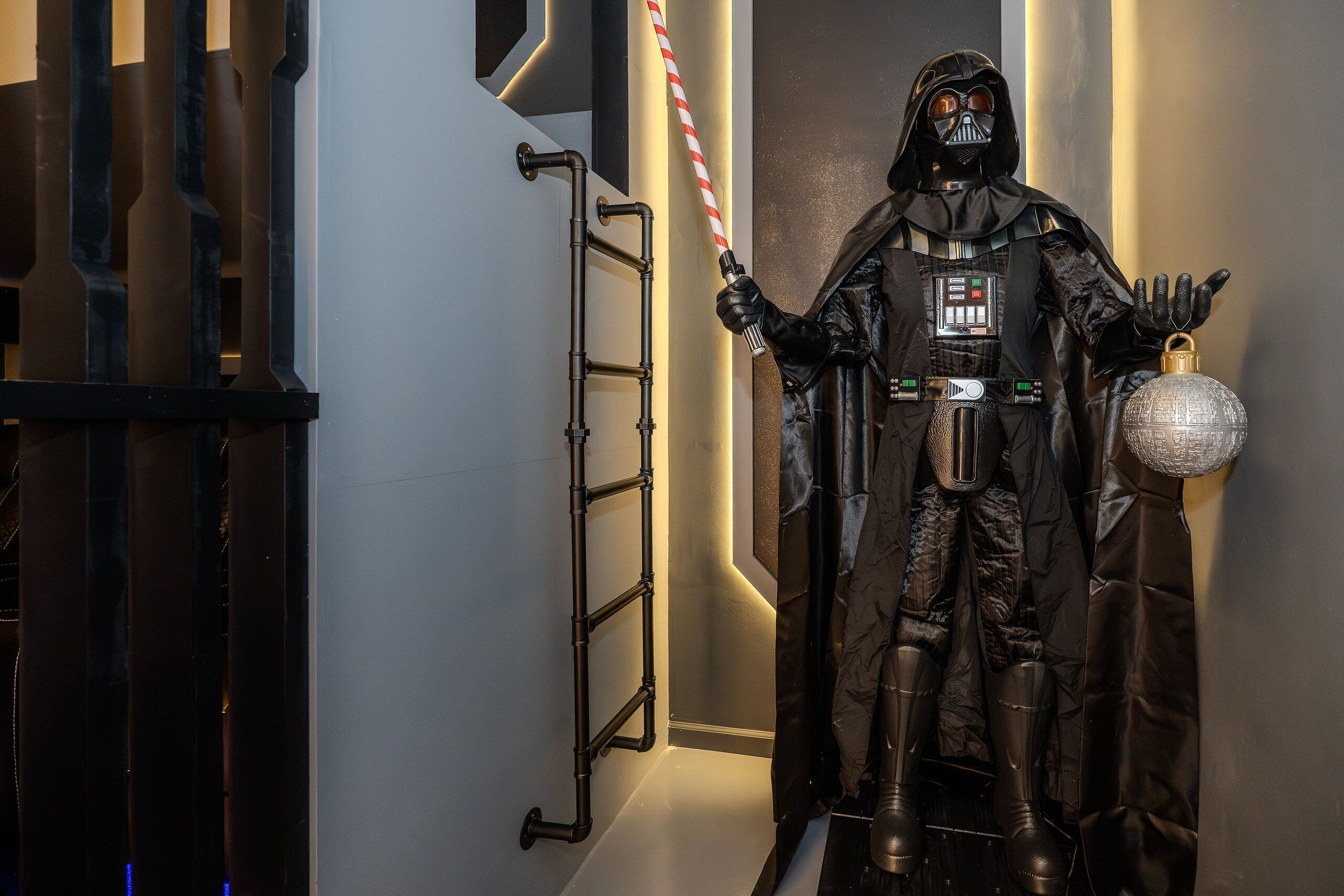 Uma estátua de Darth Vader segurando um sabre de luz em um interior moderno com tubulações aparentes e um objeto esférico decorativo.