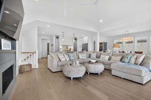 A spacious living room with a fireplace, a large sectional sofa, two round ottomans, and a kitchen in the background. - Endless Summer by Brunswick Vacation Rentals w/ Pool, Hot Tub & Walk To Beach (Ocean Isle Beach)