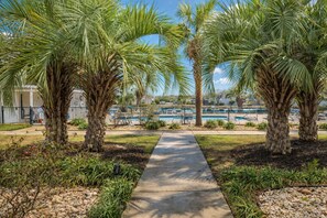 A pathway leading to a pool area with palm trees and a building in the background. - South Bay East Unit H (Myrtle Beach)