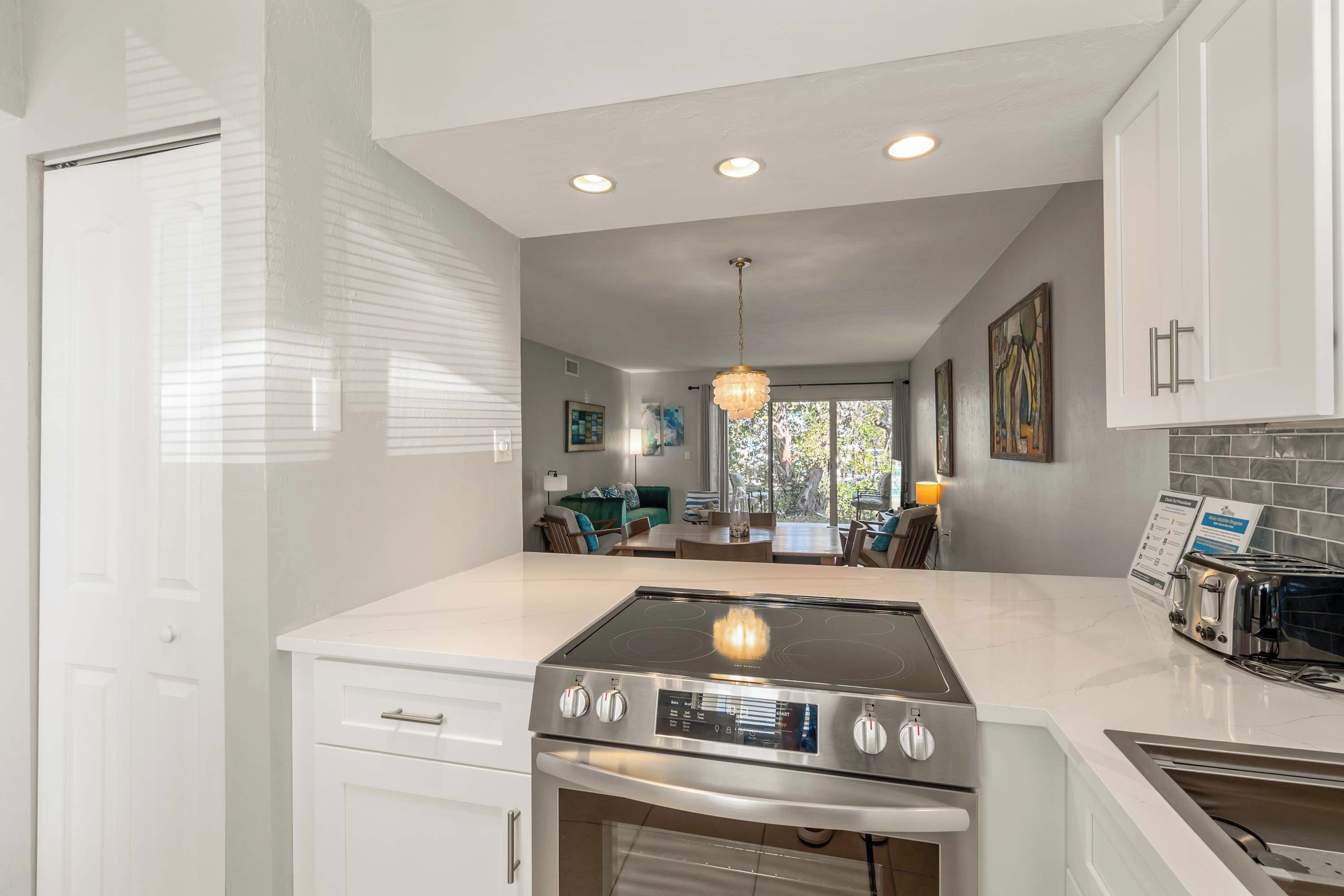 A modern kitchen with white cabinetry, a built-in oven, a stovetop, and a toaster. There is a dining area visible through the window.