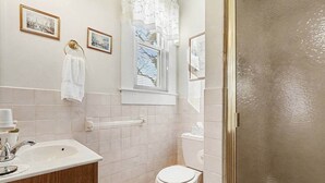 Ein Badezimmer mit Waschbecken, Waschtisch und Toilette. Zwei gerahmte Bilder hängen an der Wand, ein Handtuch hängt am Waschbecken und es gibt ein Fenster mit Jalousien.