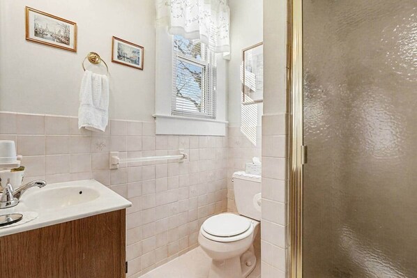 A bathroom with a white sink, vanity, and toilet. There are two framed pictures on the wall, a towel hanging on the sink, and a window with blinds.