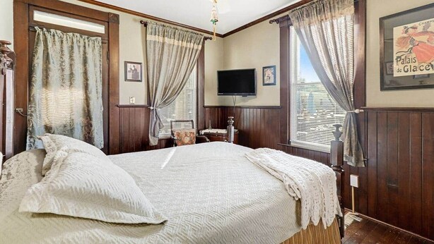 Ein Schlafzimmer mit einem großen Bett, Holzwandverkleidung, einem Deckenventilator, einem Fernseher und einem Fenster mit Vorhängen.