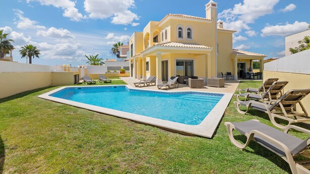 Una piscina con lettini, un edificio giallo con un balcone e palme.