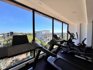 A gym with exercise equipment and large windows offering a city view. - NEW Beautiful apartment with pool view + 2 bicycles. (Tulum)