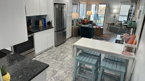 A modern kitchen with white cabinets, a stainless steel refrigerator, and a granite countertop. There is a dining area with a white table and blue chairs. - The Tiki Hut- 2 Bed, 2 Bath Oceanview Shore Drive Condo (Myrtle Beach)