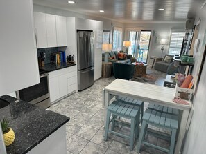 A modern kitchen with white cabinets, a stainless steel refrigerator, and a granite countertop. There is a dining area with a white table and blue chairs. - The Tiki Hut- 2 Bed, 2 Bath Oceanview Shore Drive Condo (Myrtle Beach)