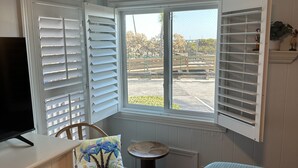 A room with a television, a chair with a floral cushion, a small round table, and a window with shutters. - The Tiki Hut- 2 Bed, 2 Bath Oceanview Shore Drive Condo (Myrtle Beach)