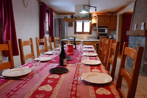 Une salle à manger avec une longue table dressée pour six personnes, des chaises en bois, une nappe rouge et une cuisine en arrière-plan.