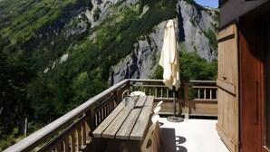 Una terraza de madera con una mesa, sillas, un banco y una sombrilla, con vistas a un paisaje montañoso.