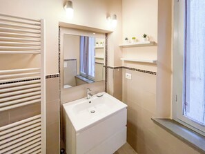 A bathroom with a white sink, mirror, and towel rack. There are two wall-mounted lights and a window with a view. - Michelangelo Heart of Tuscany (Peccioli)