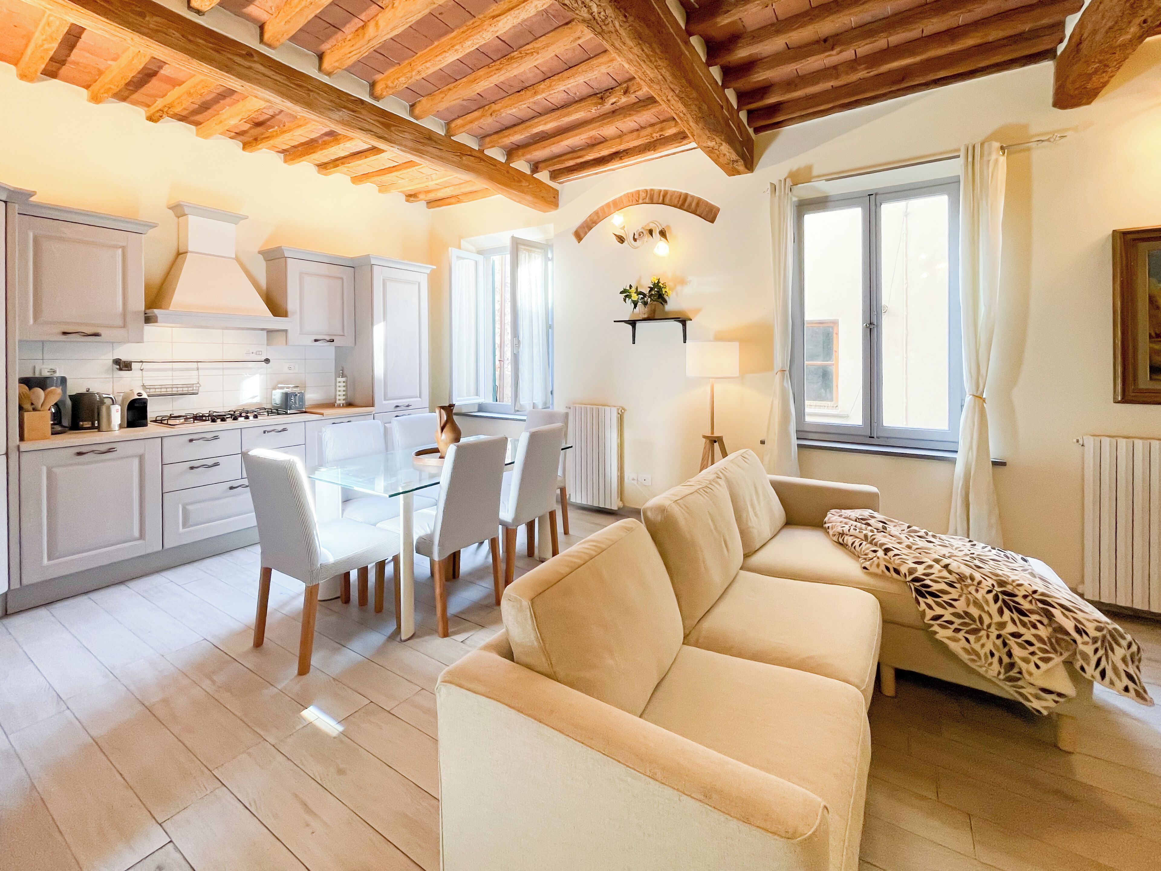 A modern kitchen with white cabinets, a dining table with chairs, and a beige sofa in a bright living area with wooden beams on the ceiling.