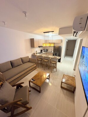 A modern living room with a sofa, coffee table, and chairs. A kitchen is visible in the background with a dining area.