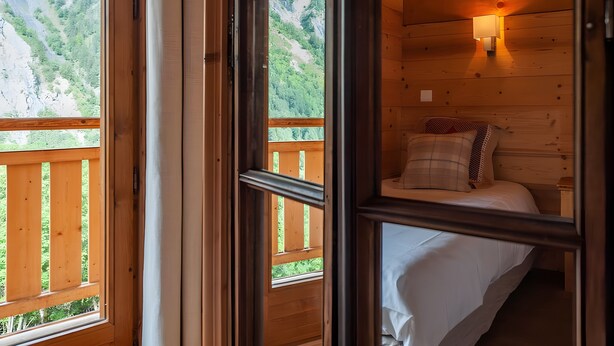 A wooden cabin room with a bed, a nightstand, a wall-mounted lamp, and a window with a view of a mountainous landscape. - Chalet “Eden” with Balcony and Wi-Fi (Les Belleville)