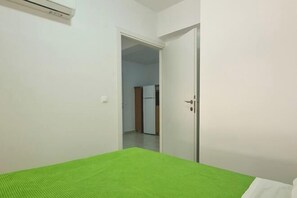 Ein kleiner, sauberer Raum mit einem Bett, einer Tür, die in einen anderen Raum führt, und einer weißen Wand.
