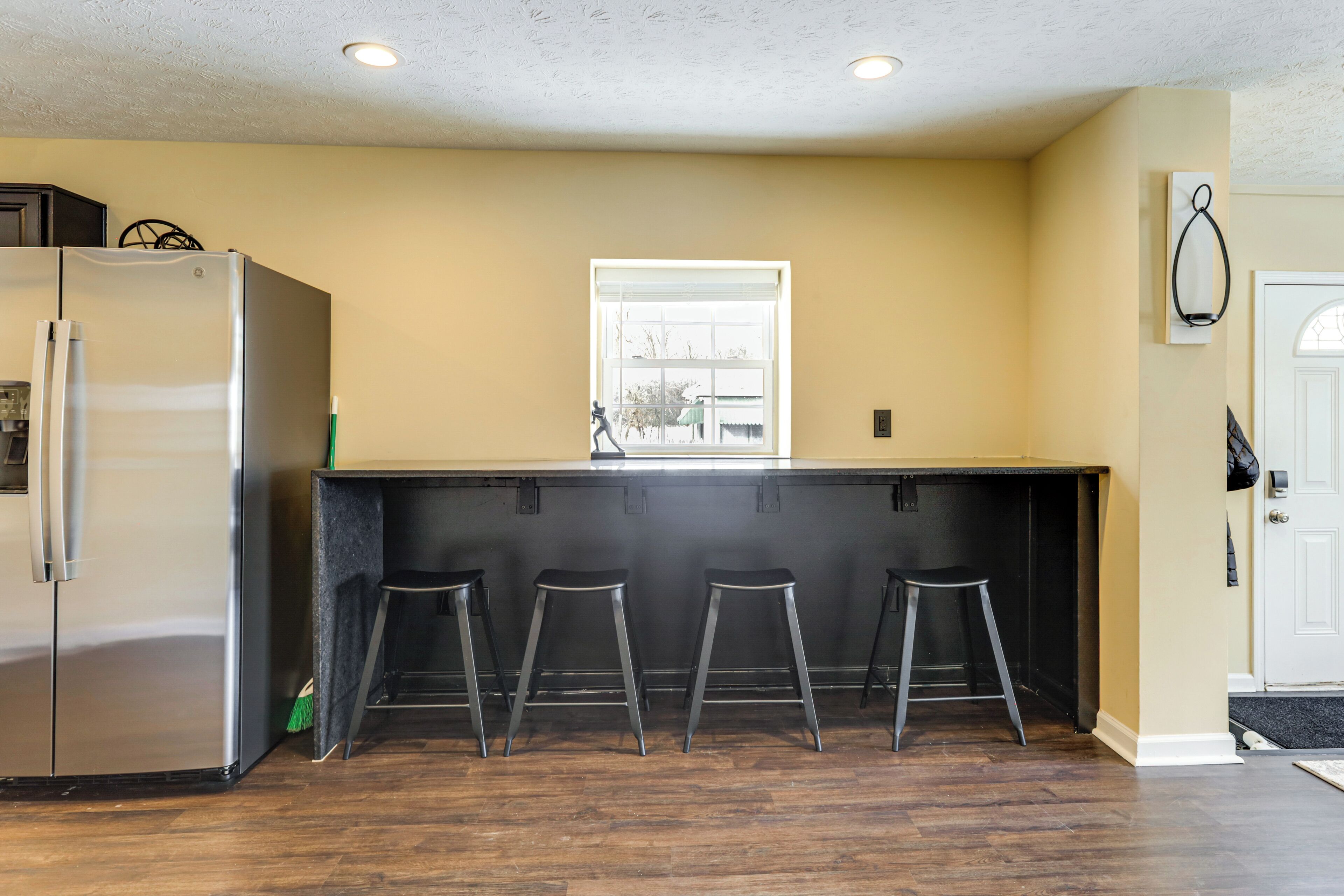 A kitchen with a stainless steel refrigerator, a window above the counter, and three stools.