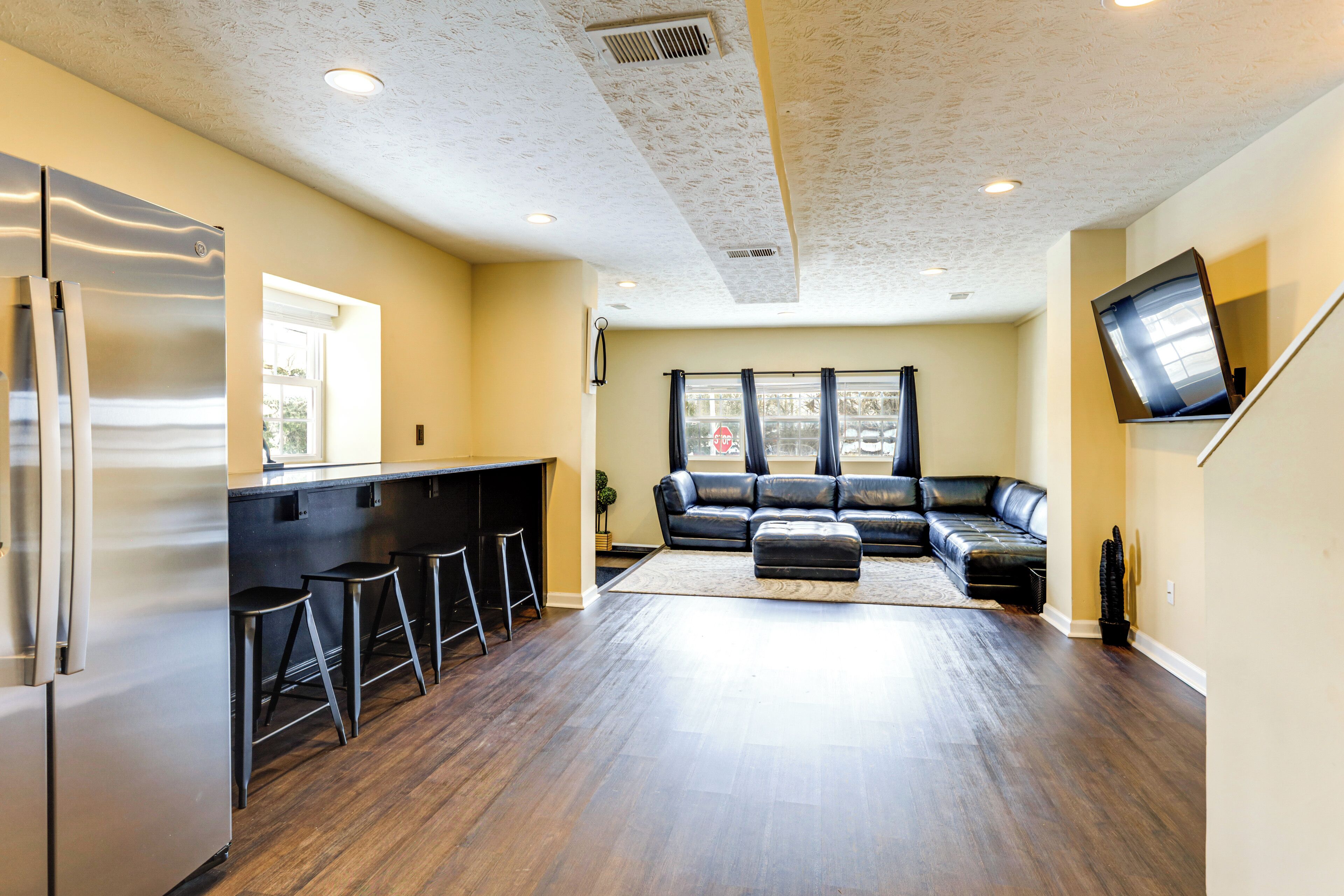 A modern kitchen with a stainless steel refrigerator, a bar area with stools, a black leather sofa, and a flat-screen TV mounted on the wall.