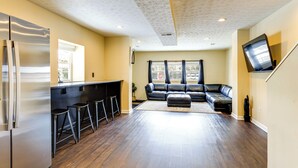 A modern kitchen with a stainless steel refrigerator, a bar area with stools, a black leather sofa, and a flat-screen TV mounted on the wall. - Townhome: Bunk Room for 6, Dry Bar Lounge Area (Cincinnati)