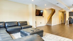 A spacious living room with a sectional sofa, ottoman, and patterned rug. A staircase leads to a kitchen area. - Townhome: Bunk Room for 6, Dry Bar Lounge Area (Cincinnati)